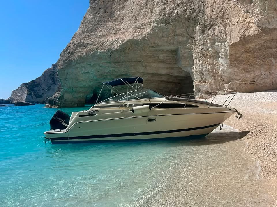 ΝΙΚΟΛΑΟΣ, the cream MareWaves cabin cruiser with its blue bimini up, resting bow-first on a pebble beach beneath an overhanging white cliff, turquoise shallows in front of it