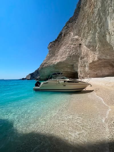 ΝΙΚΟΛΑΟΣ pulled up on the pebble shore of an empty cove under a high cliff, clear turquoise shallows in front