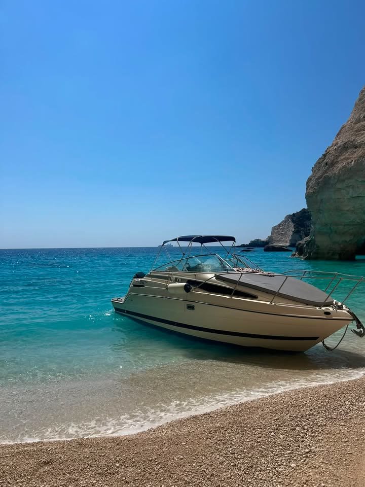ΝΙΚΟΛΑΟΣ nosed onto a sandy shoreline with the bimini up and the bow rail over the beach, open blue sea and a rock headland behind