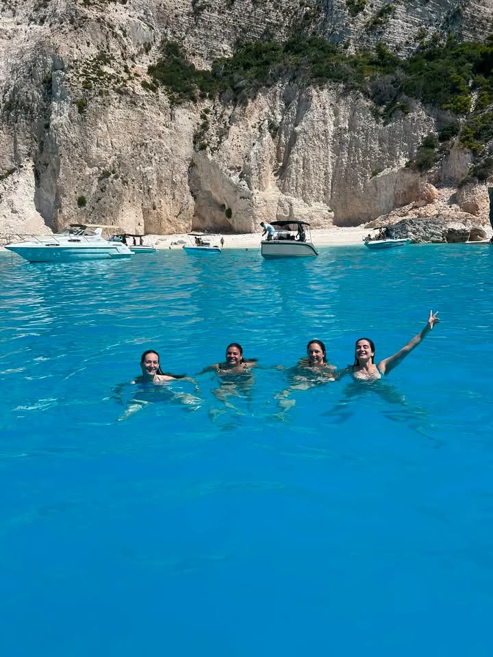 Four guests swimming in vivid blue water below the white cliffs of Zakynthos, with the MareWaves boat anchored behind them
