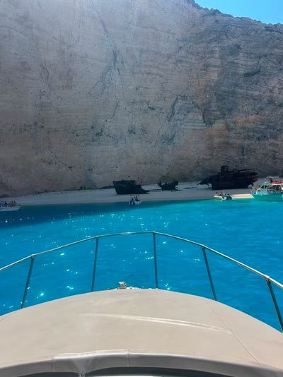 The rusted Navagio shipwreck on its white sand beach beneath towering cliffs, seen from the bow of the boat