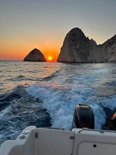 The boat's wake glowing orange at sunset with the Mizithres rocks and the setting sun behind